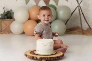 Baby boy wearing a tan outfit with a birthday cake and balloons in neutrals and shades of green