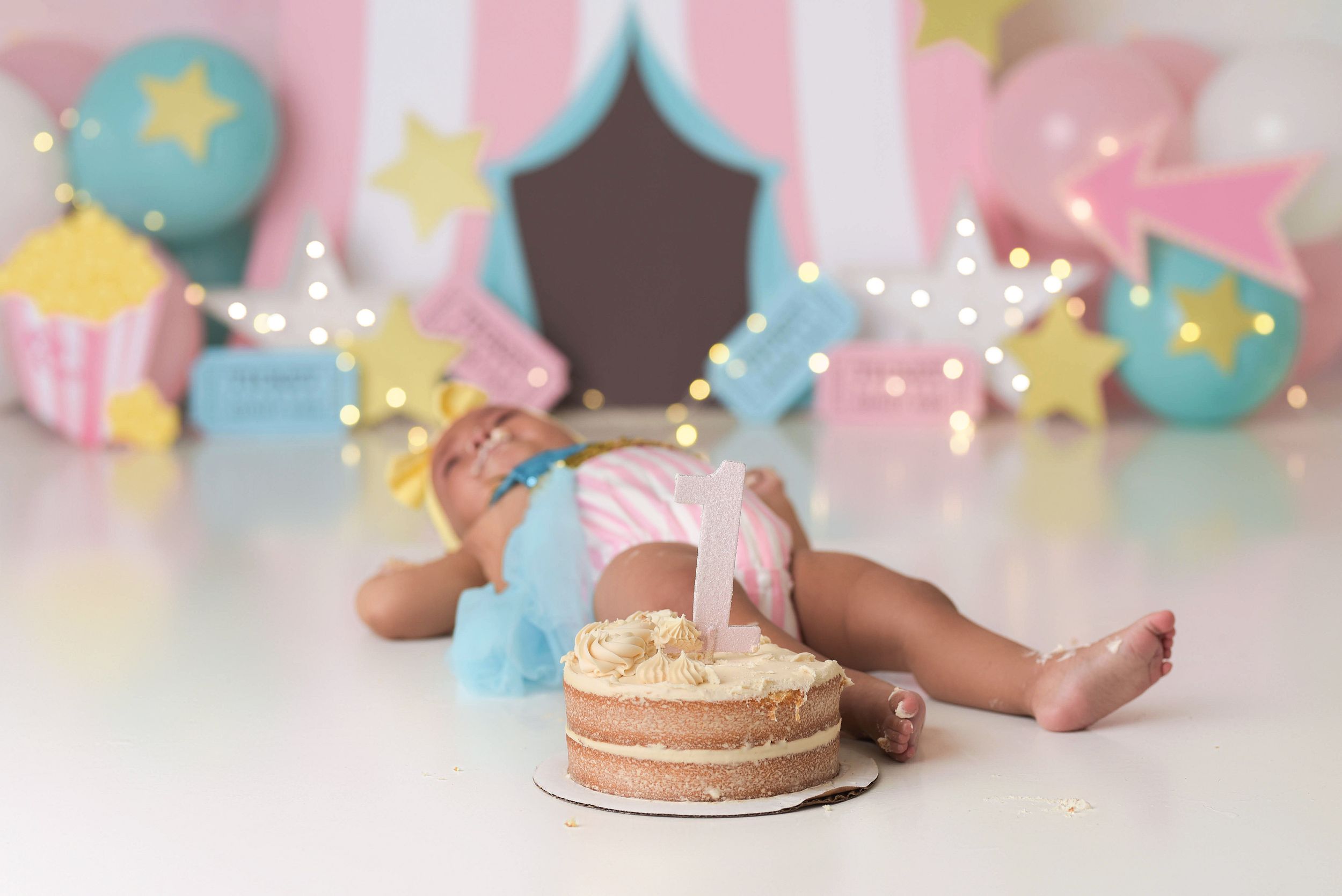 Baby girl laying on her back by her cake after eating too much birthday cake. Her setup is a fun pastel circus theme