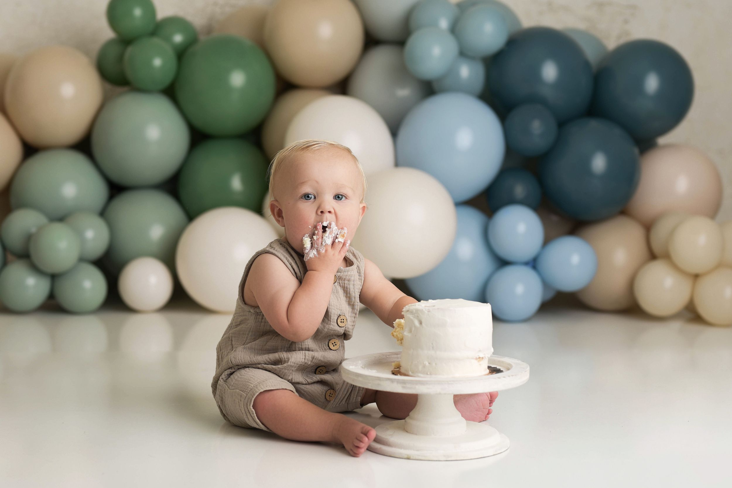 Baby boy smashing his first birthday cake in front of a wall of blue and green balloons