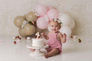 Baby girl in front of a simple rustic strawberry first birthday themed setup with a small balloon garland in tan and pink.