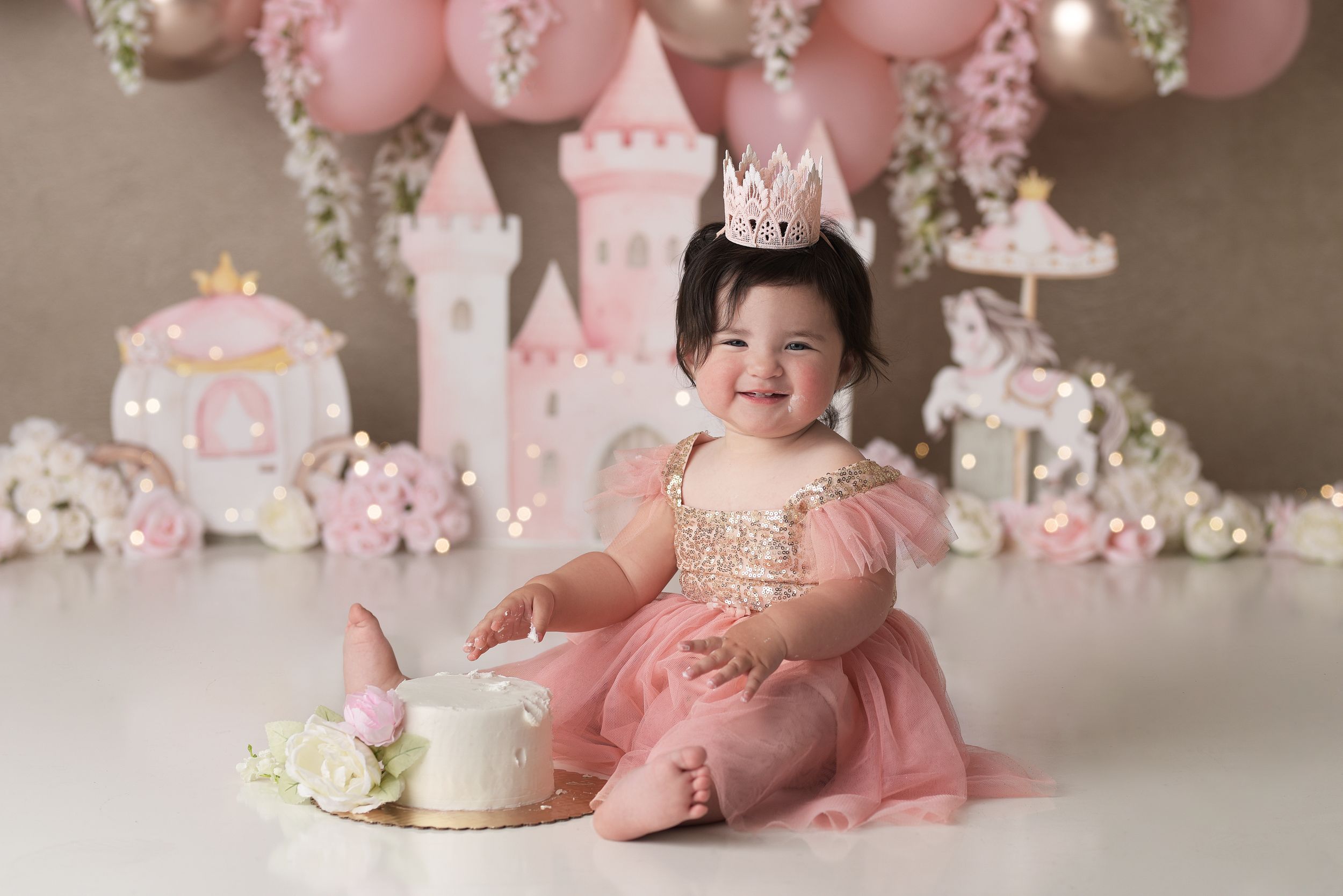 Baby girl with a pink and gold castle princess backdrop with her birthday cake, wearing a pink and gold sparkly dress and pink crown.
