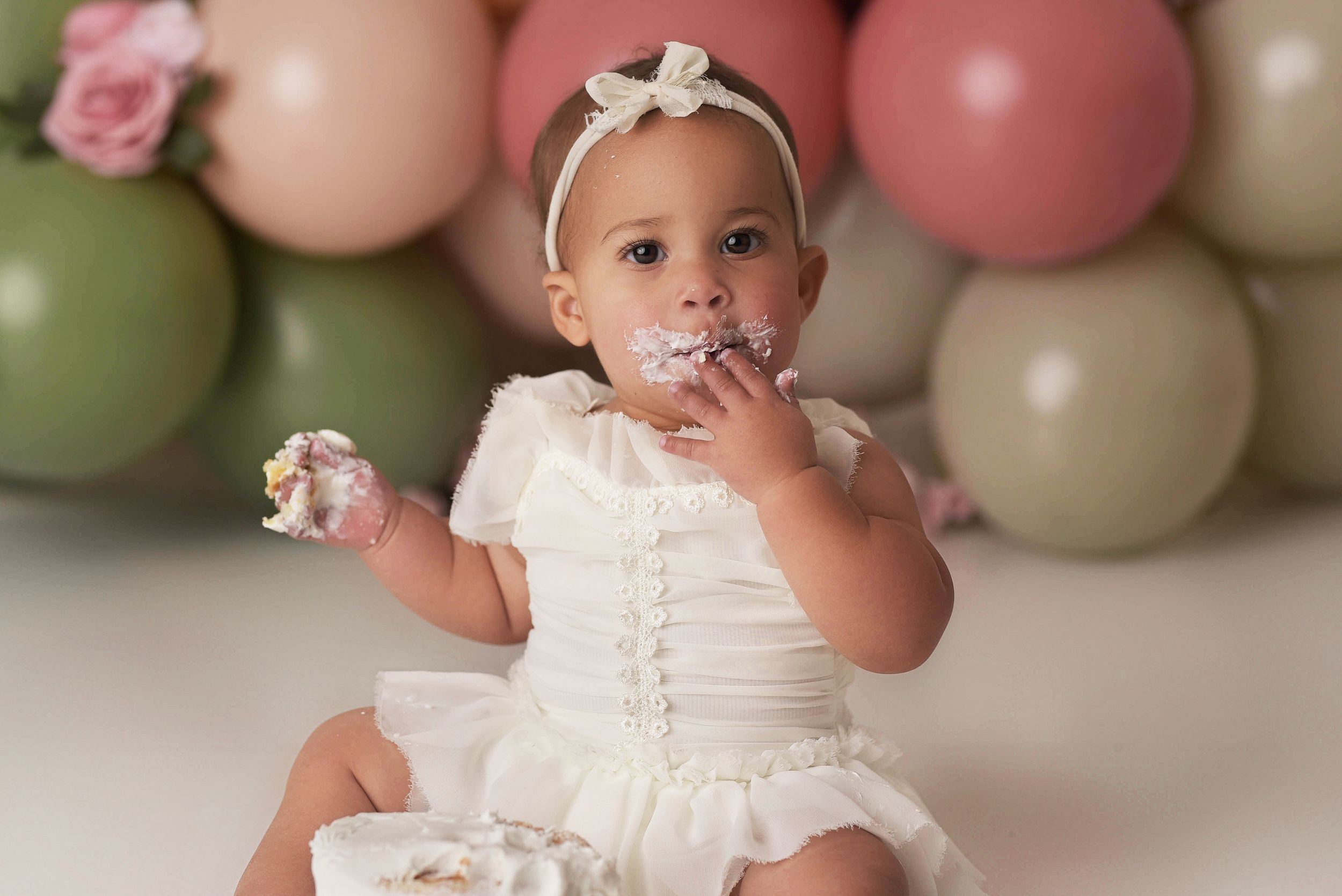 Baby girl eating a handful of birthday cake with a balloon backdrop in shades of green and pink