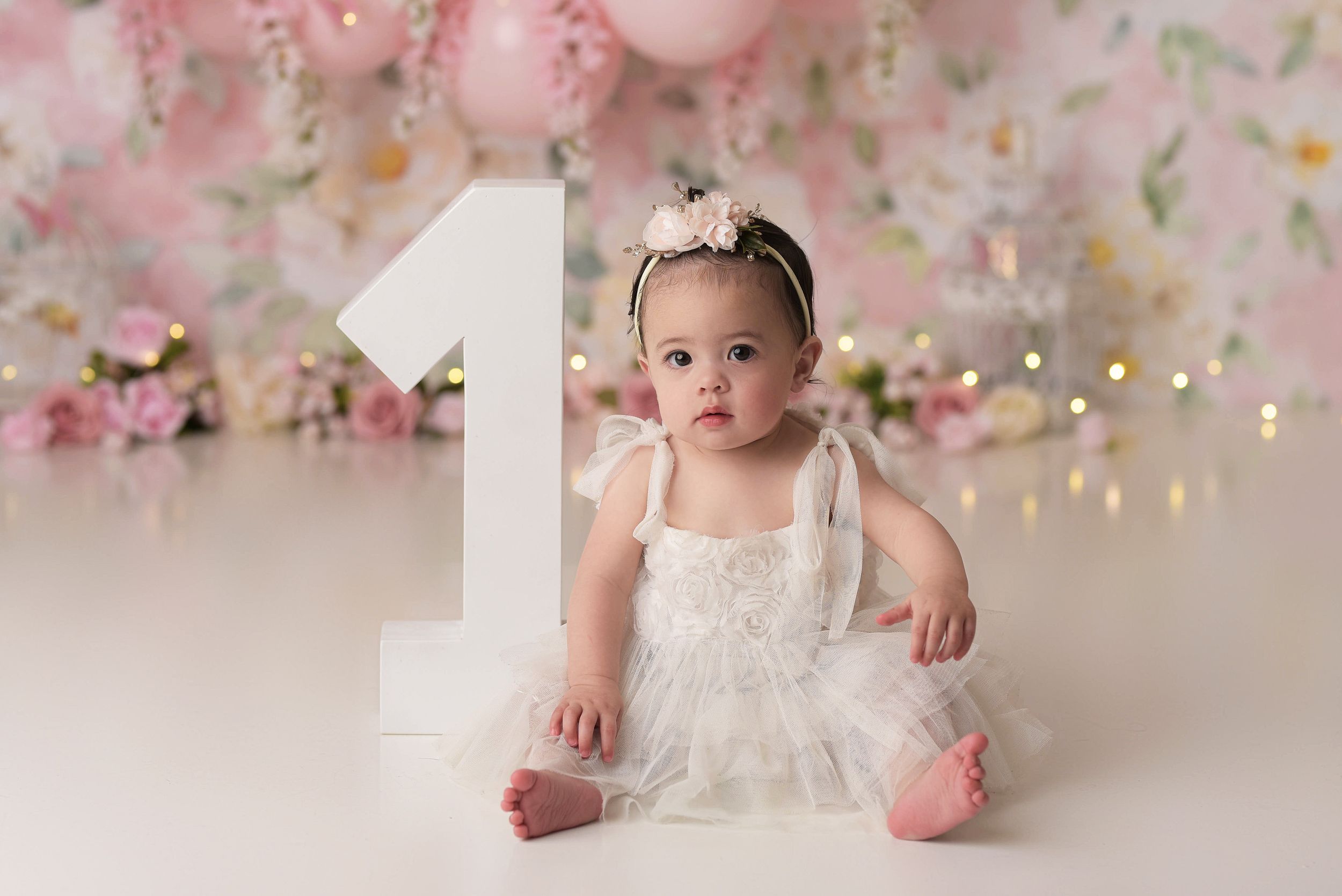 Baby girl in white with a pink floral backdrop sitting with an oversized number one
