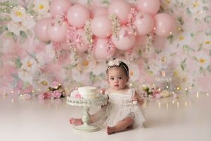 Baby girl wearing white with her white birthday cake, in front of a light pink and white floral backdrop and lots of light pink balloons