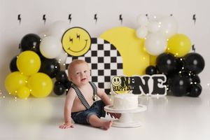 Baby boy with a one happy dude cake smash setup in bright yellow and black & white balloons and a checkered patterned arch.