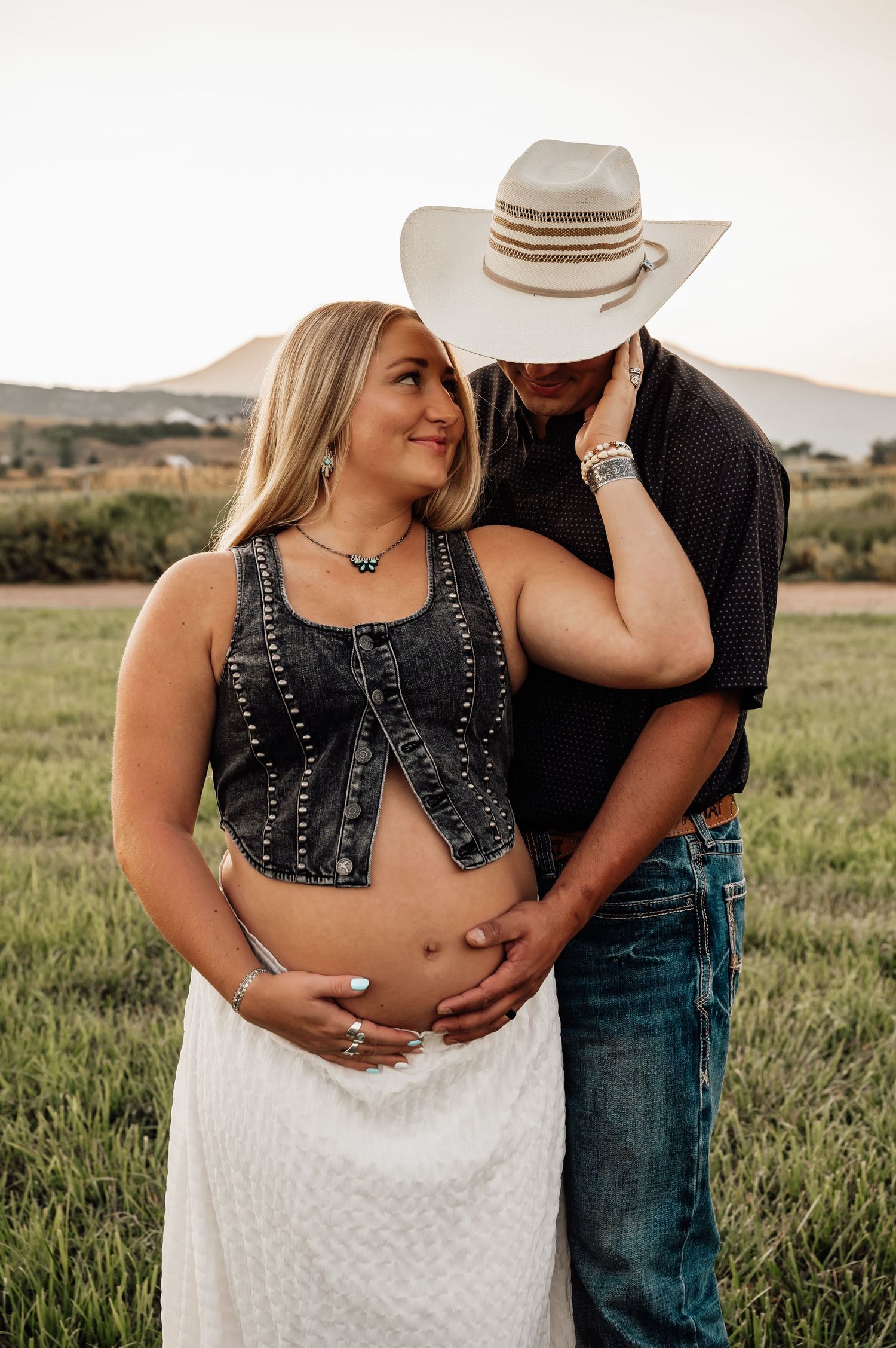 Couple in western wear embracing in a grassy field at sunset.