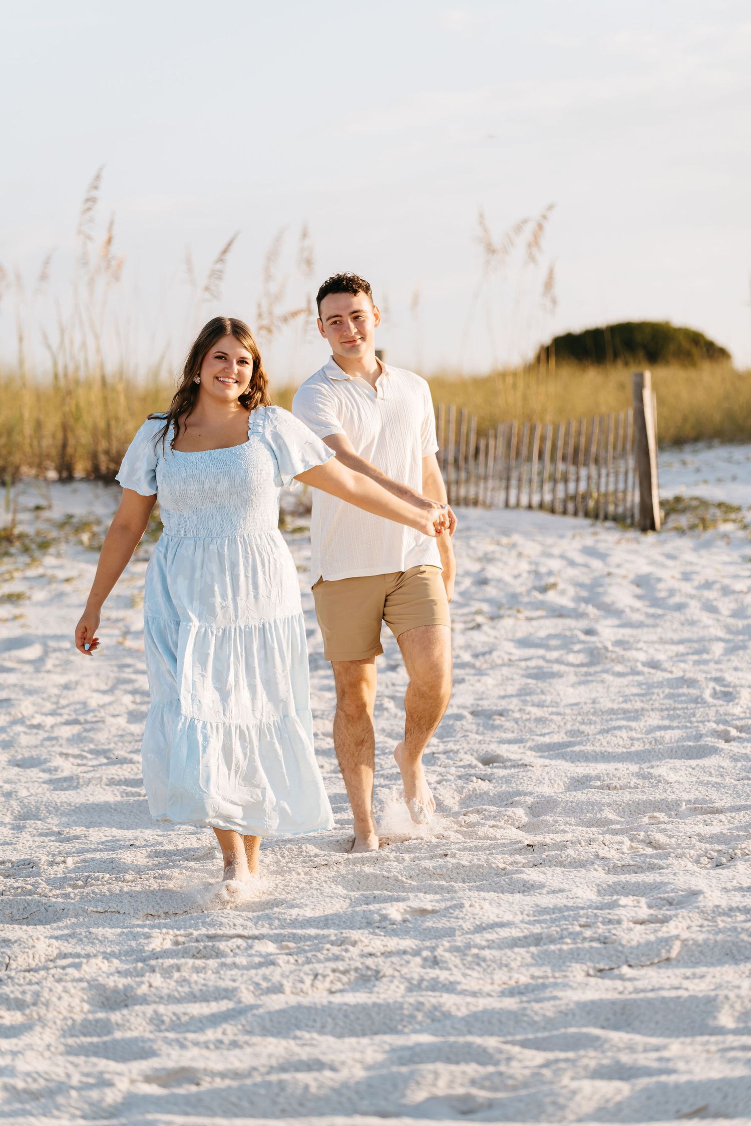 A couple walks together on pristine white sand beach at sunset with sea oats in the background.