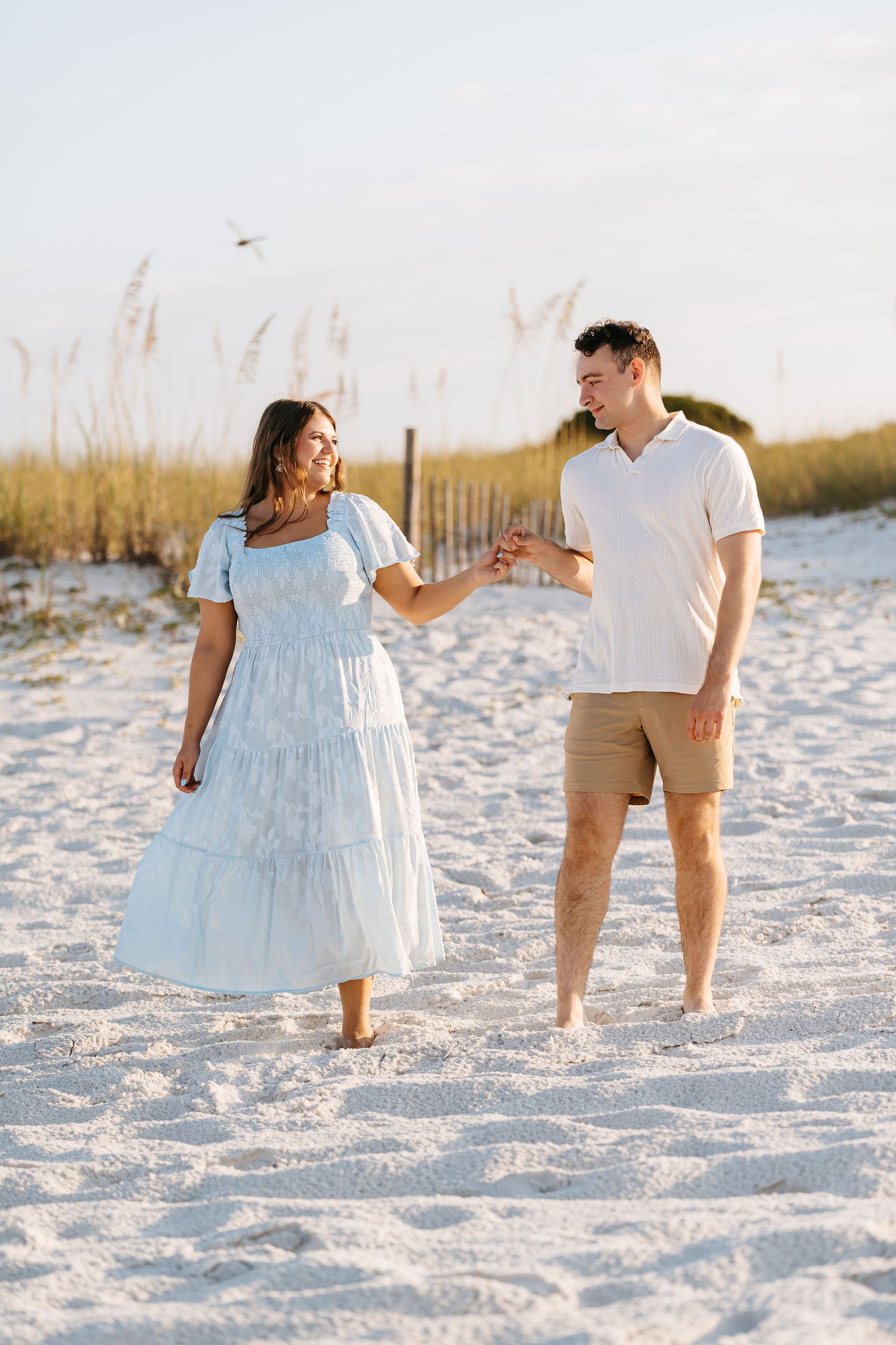 A couple walks barefoot along a sandy beach at sunset, with the woman wearing a flowing white dress.