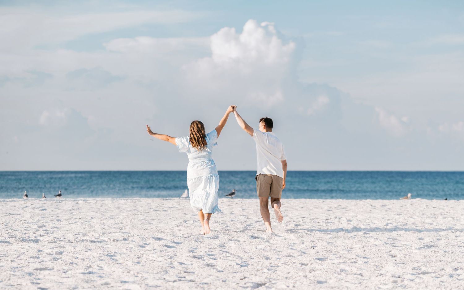 Couple in white outfits walking along a pristine white sandy beach with turquoise ocean waters in the background.