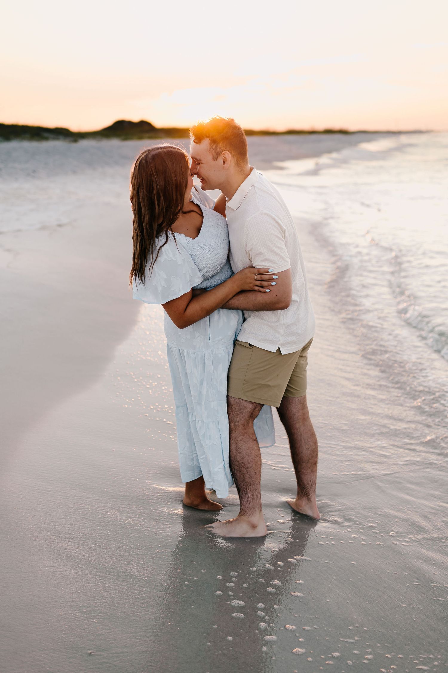 A couple shares a romantic moment while standing in the shallow ocean waves at sunset on a sandy beach.