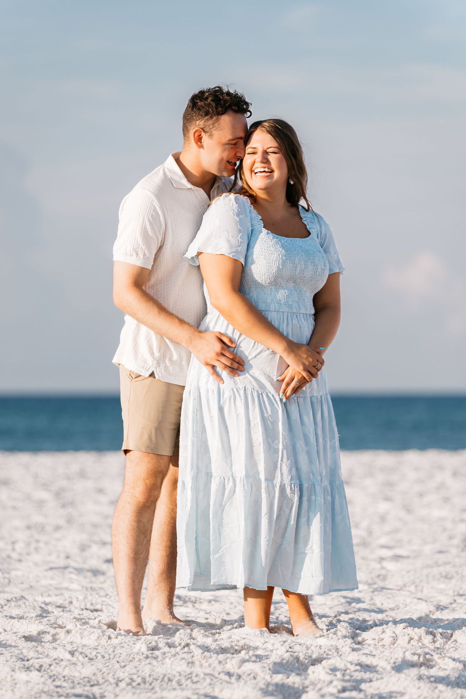 A couple in white outfits embraces on a pristine white sand beach with ocean waves in the background.