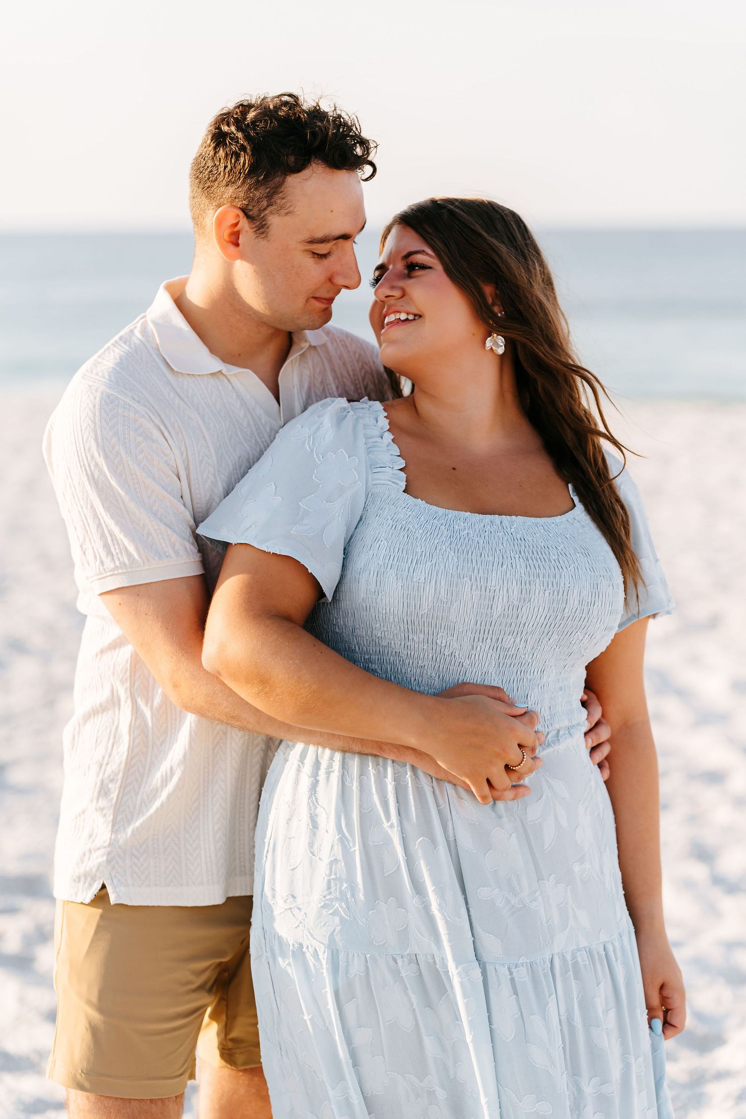 A romantic couple embraces on a white sandy beach wearing coordinated light colored outfits. Local Couples Photographer for candid photos.