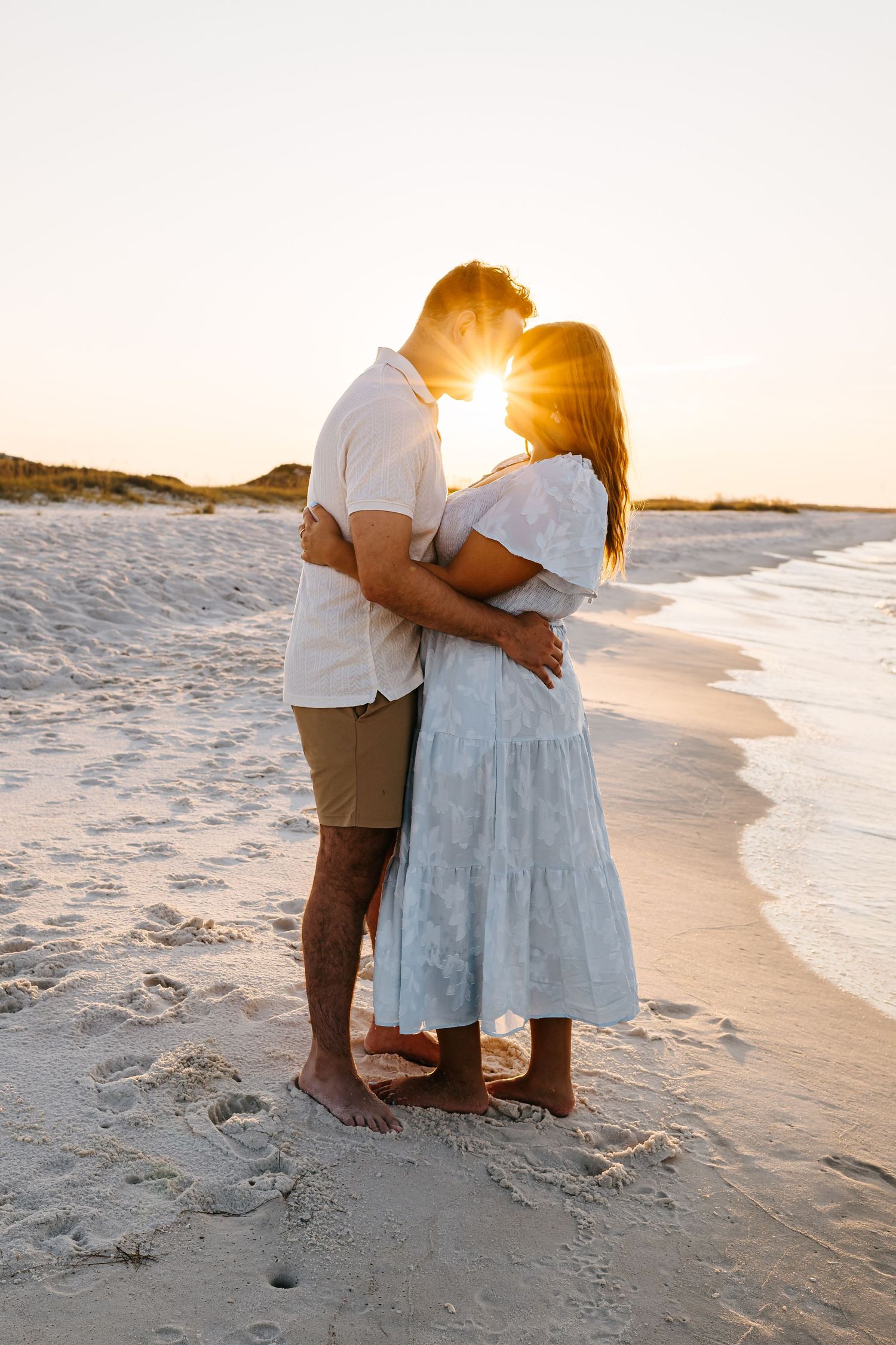 A couple embraces on a sandy beach at sunset as golden sunlight streams through the scene.