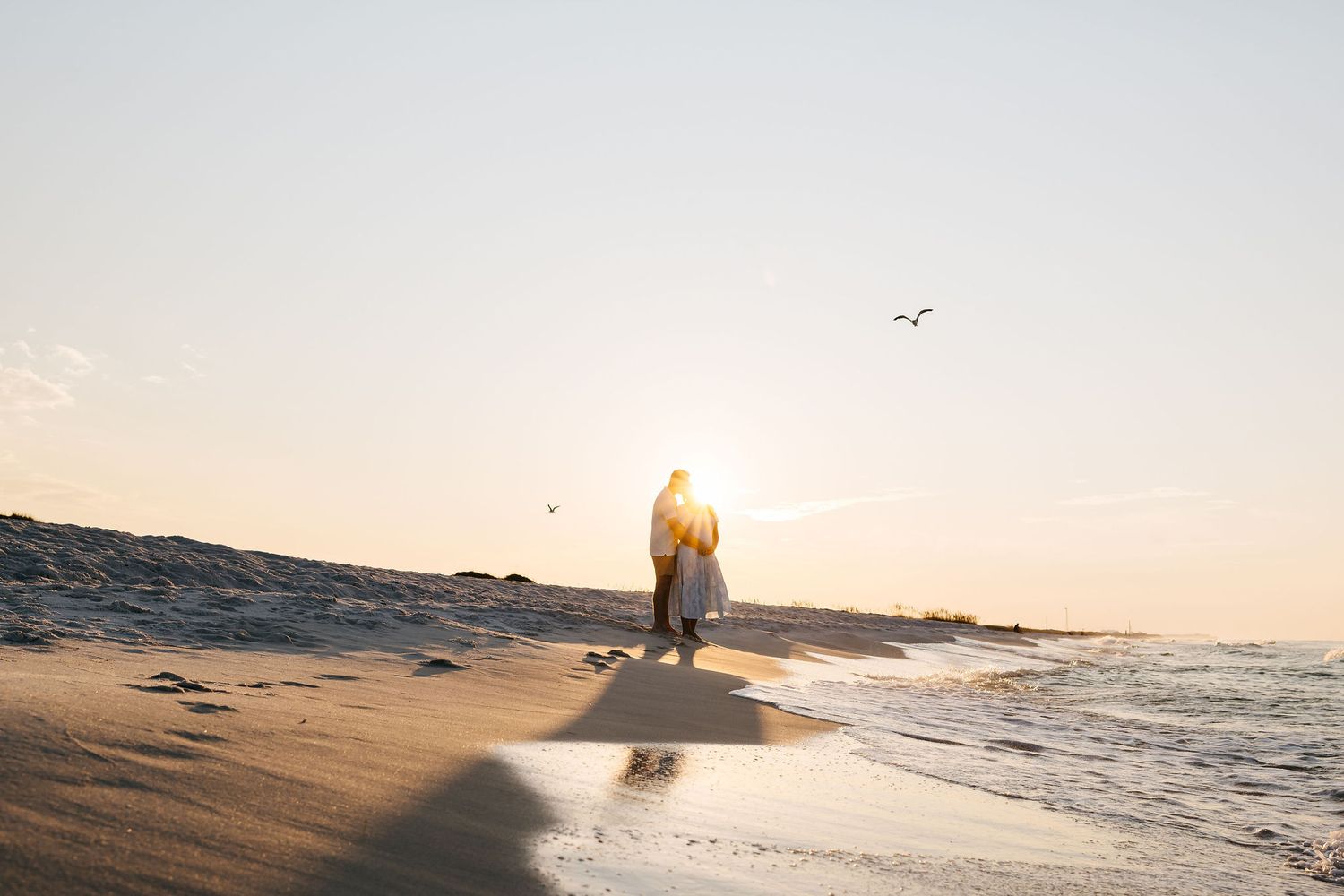 A silhouetted figure stands on a sandy beach at sunset with ocean waves rolling in and a bird flying overhead.