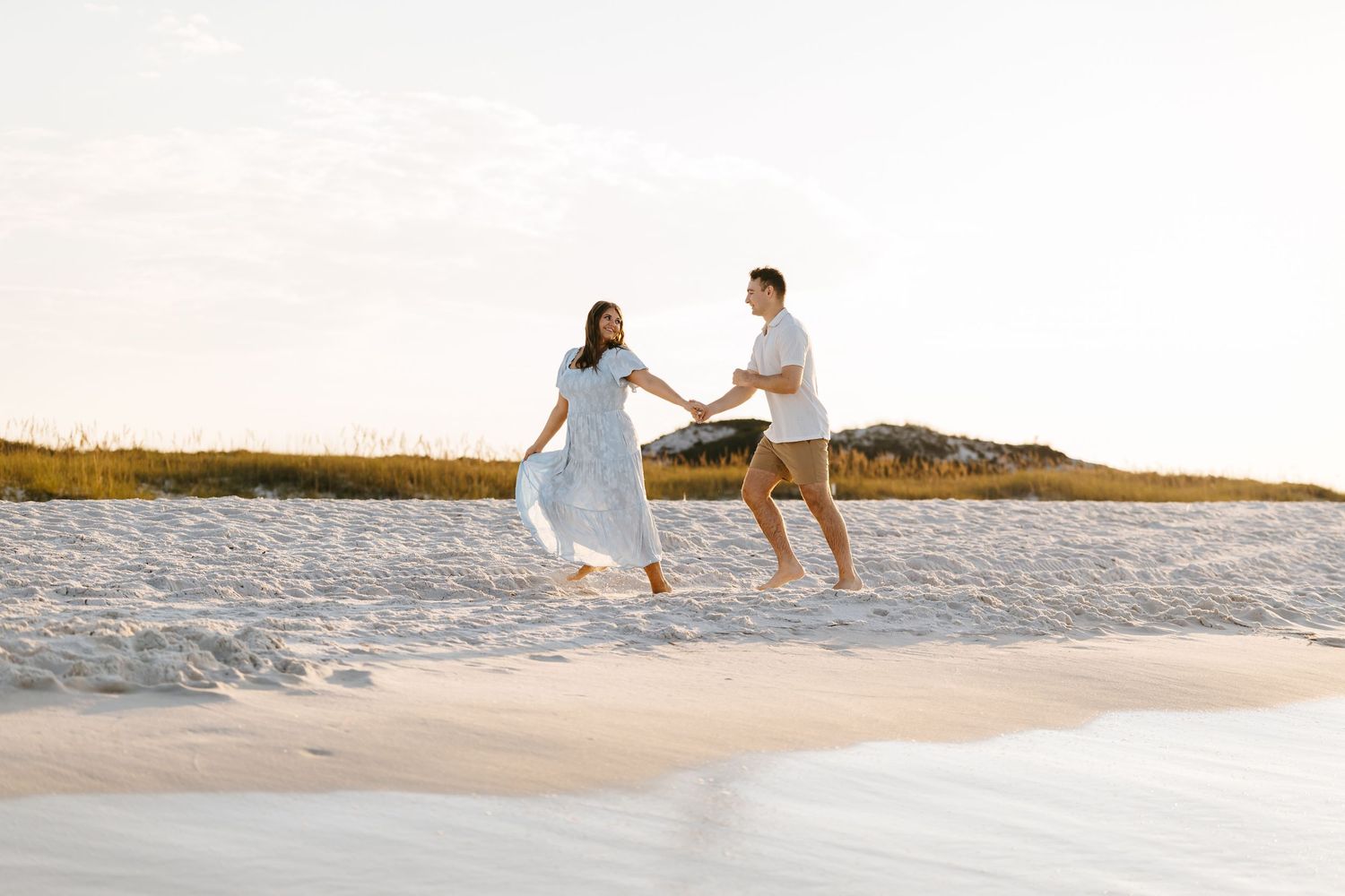 A couple in white outfits walks along a scenic beach at sunset holding hands and enjoying a romantic moment.
