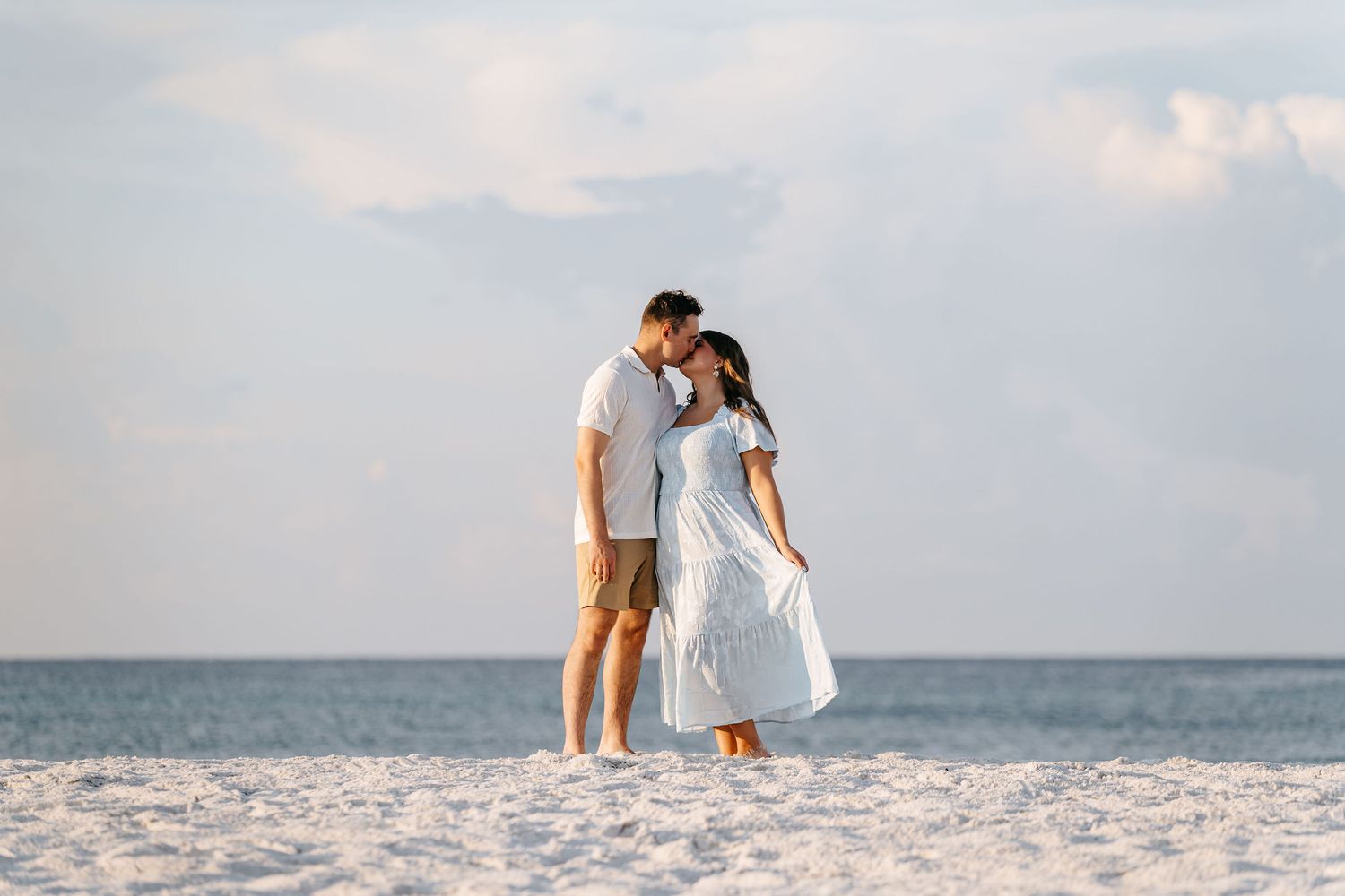 A couple embraces and kisses on a white sandy beach with the ocean and cloudy sky in the background.