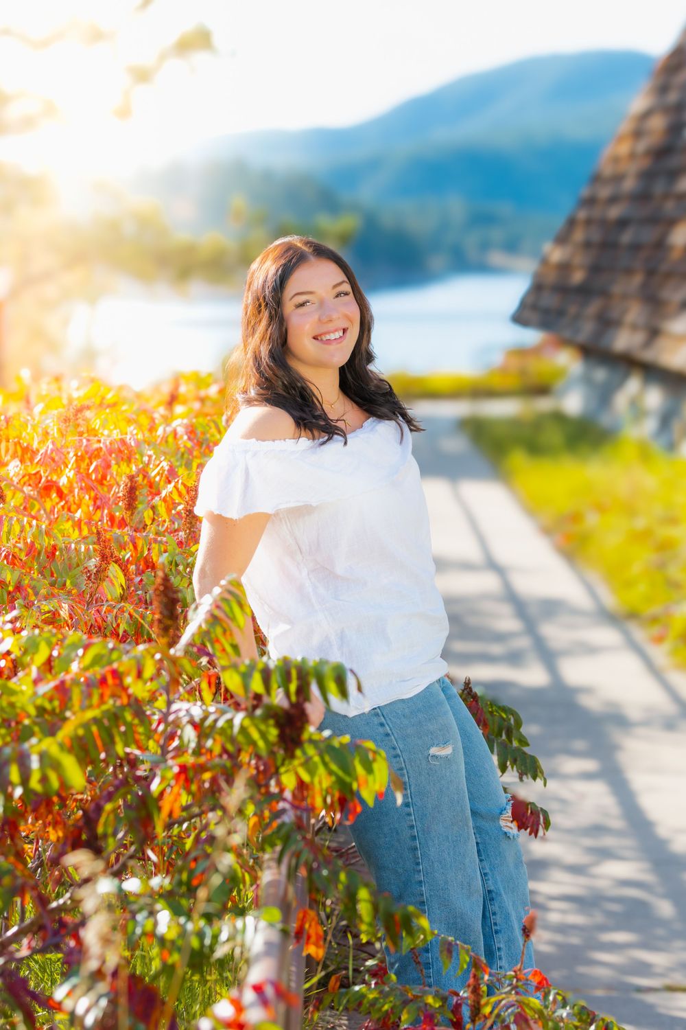 A person in a white top and jeans poses outdoors against vibrant autumn foliage and a scenic mountain backdrop.
