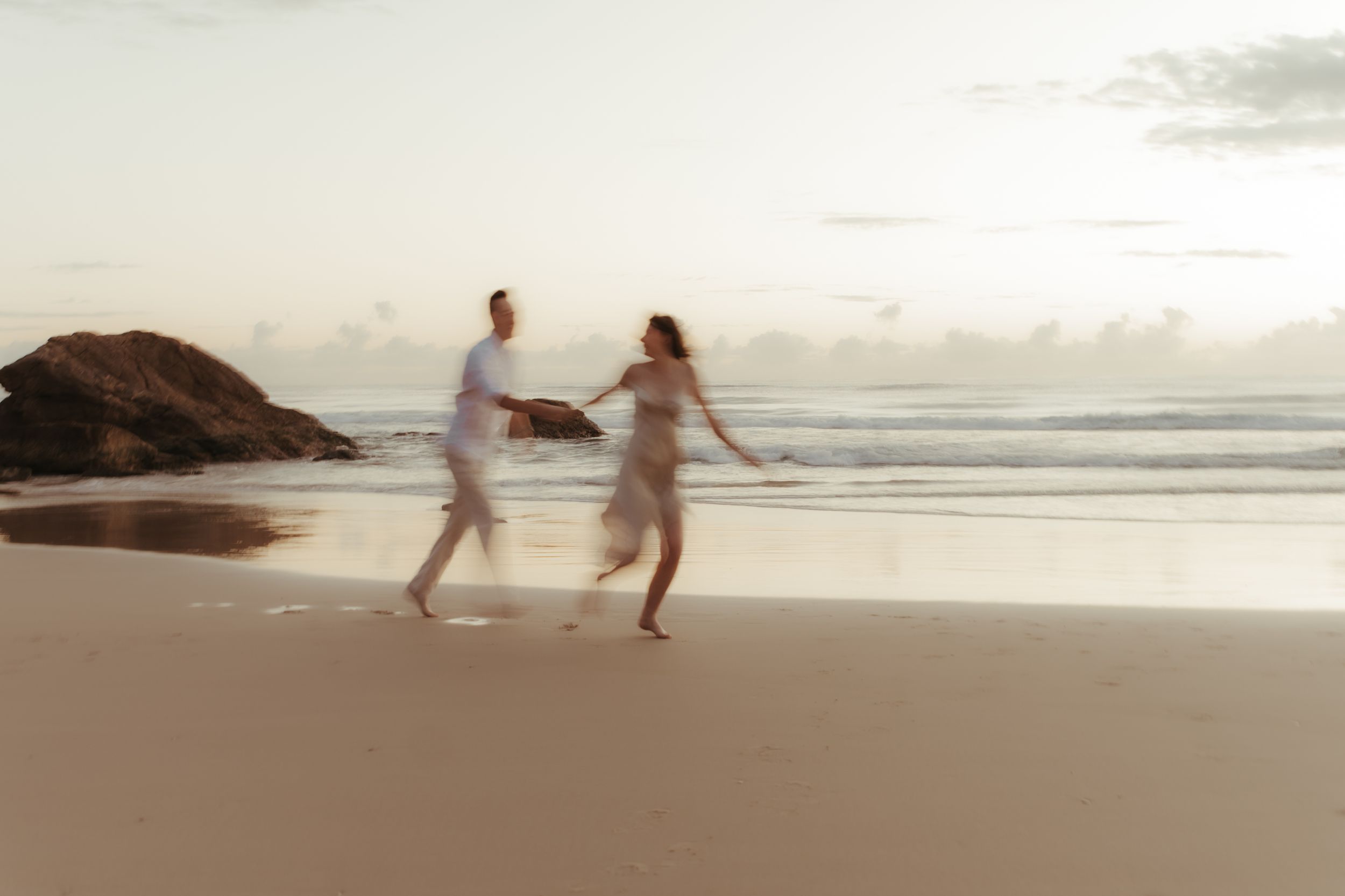 People running and playing together on a sandy beach at sunset with ocean waves and rocky cliffs in the background.
