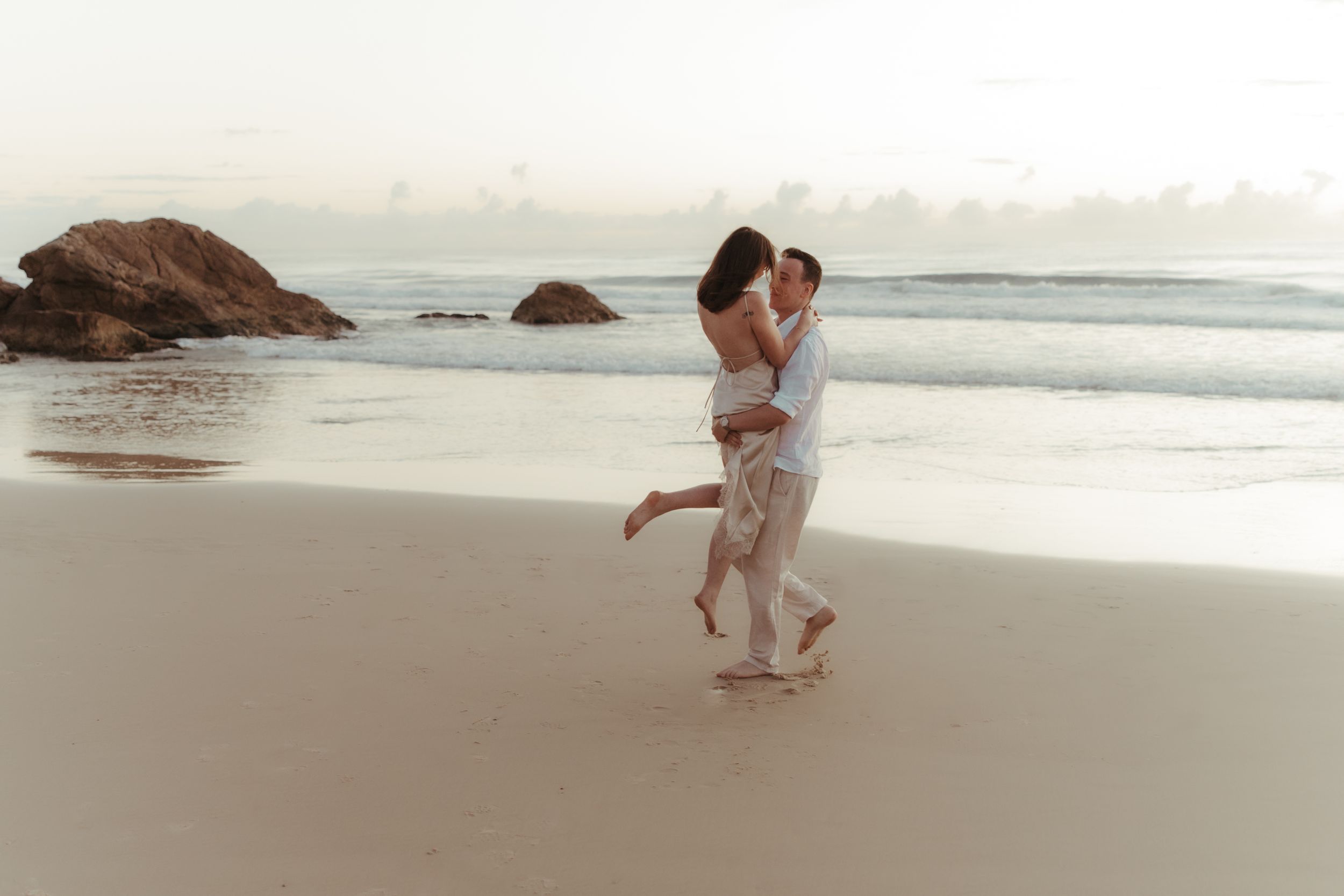 A romantic couple embraces while standing on a misty beach with rocky outcrops in the background.