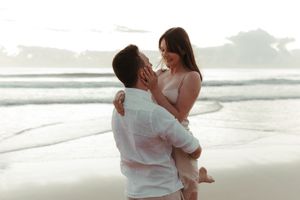 A romantic couple in white outfits embraces on a misty beach at sunset.