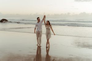 Couple holding hands and walking together on a misty beach at sunset with waves crashing in the background.