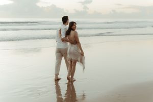 Couple in white outfits walking together along a misty beach shoreline at sunset.