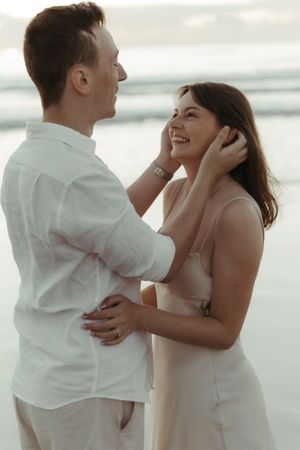 A couple in white outfits shares intimate moments on a beach in a series of romantic engagement photos.