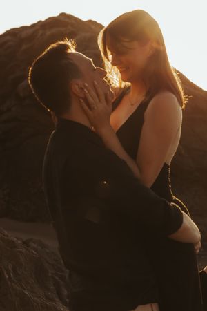 Romantic sunset photos of couple embracing on rocky beach with golden backlighting.