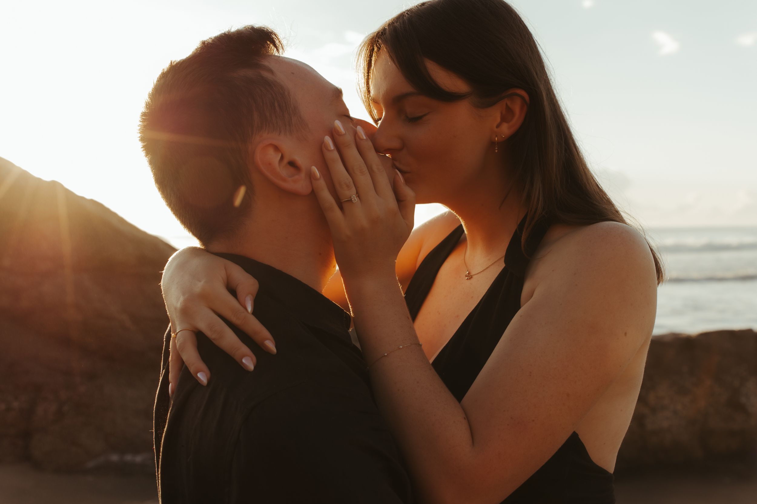 A couple shares a romantic kiss on the beach at sunset while waves crash behind them.