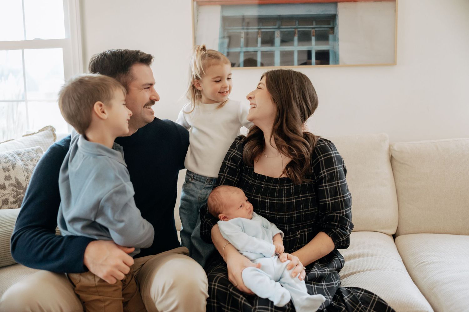 Happy family of five sitting on white couch, parents smiling with two kids and a newborn baby.