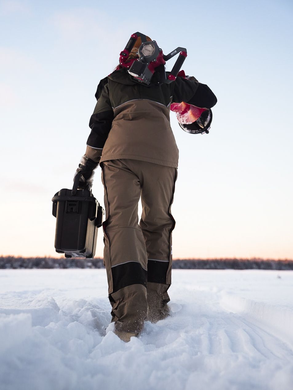 Ice fisher kneeling on frozen lake surface at sunset while checking fishing equipment.