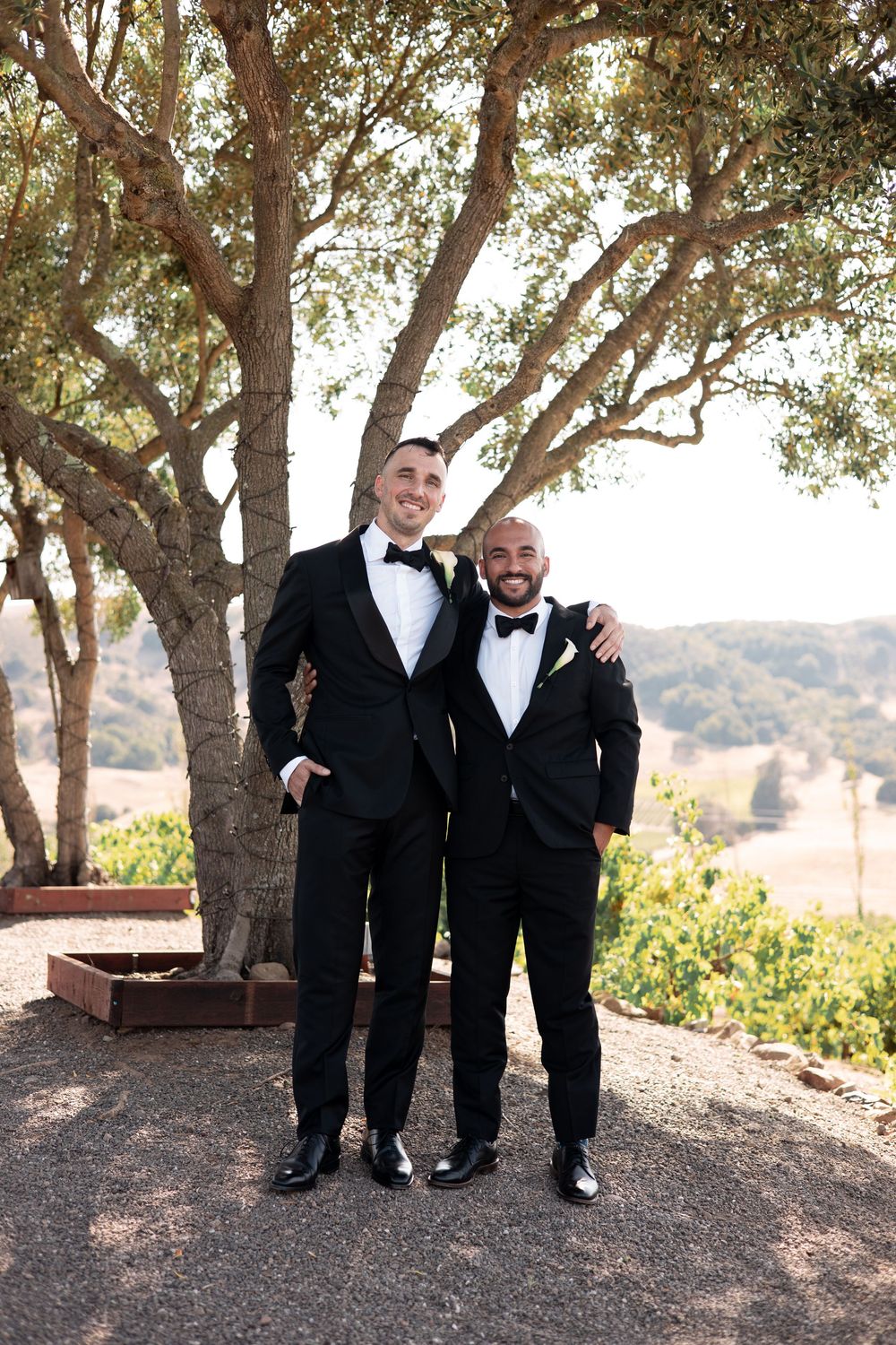 Two people in formal black tuxedos share candid moments together under trees in an outdoor setting.