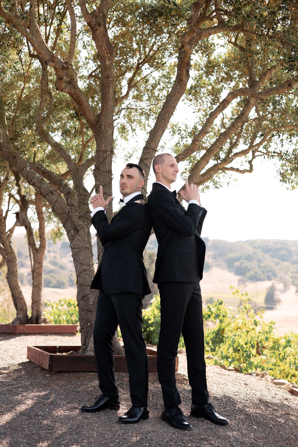 Wedding party members in black suits striking animated poses beneath a grove of trees.
