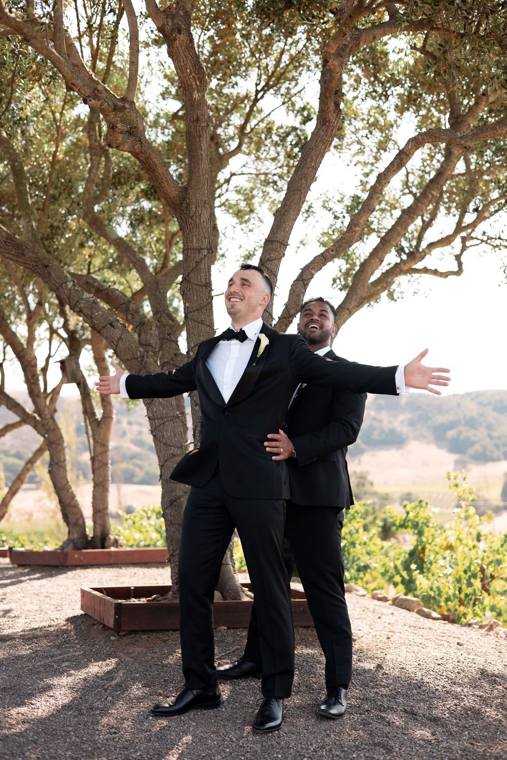 People in formal attire making dramatic gestures and posing playfully under a canopy of trees.