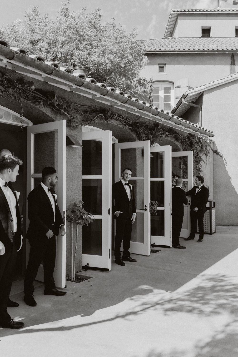 A black and white photo shows groomsmen lined up along a covered walkway with white doors.