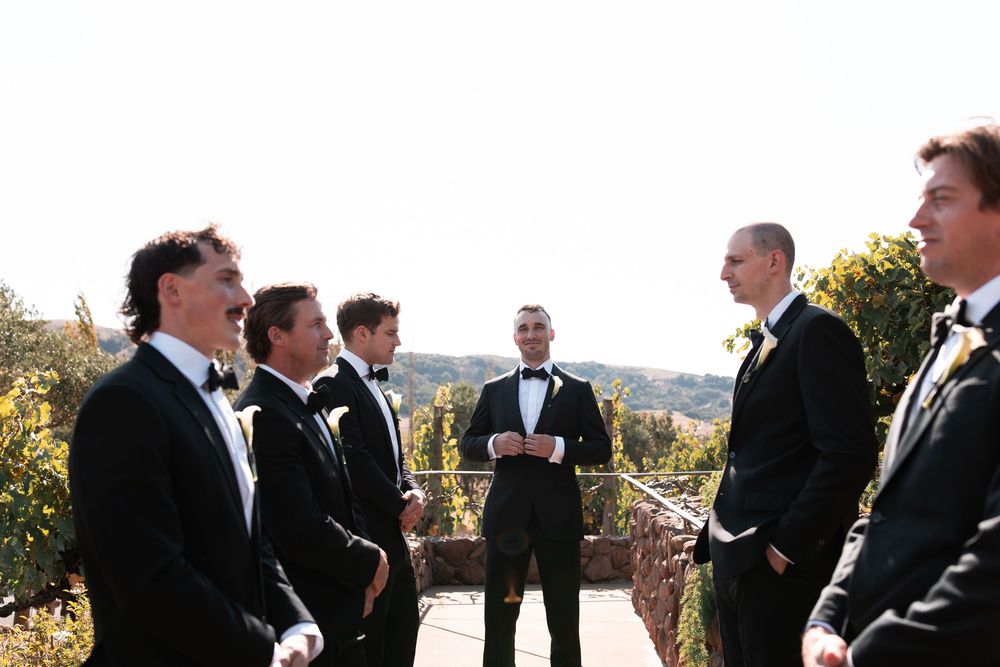 Group of formally dressed people gathered outdoors for wedding photos under natural lighting.