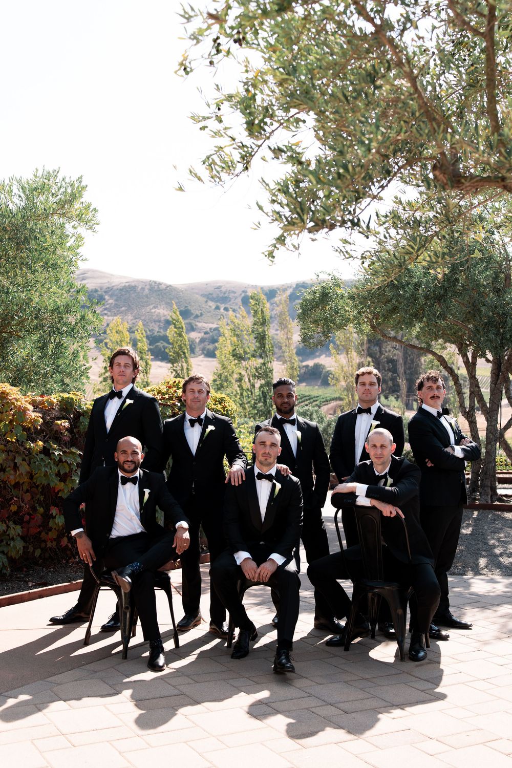 A group in formal black tuxedos sits on a stone wall outdoors with trees and mountains in the background.