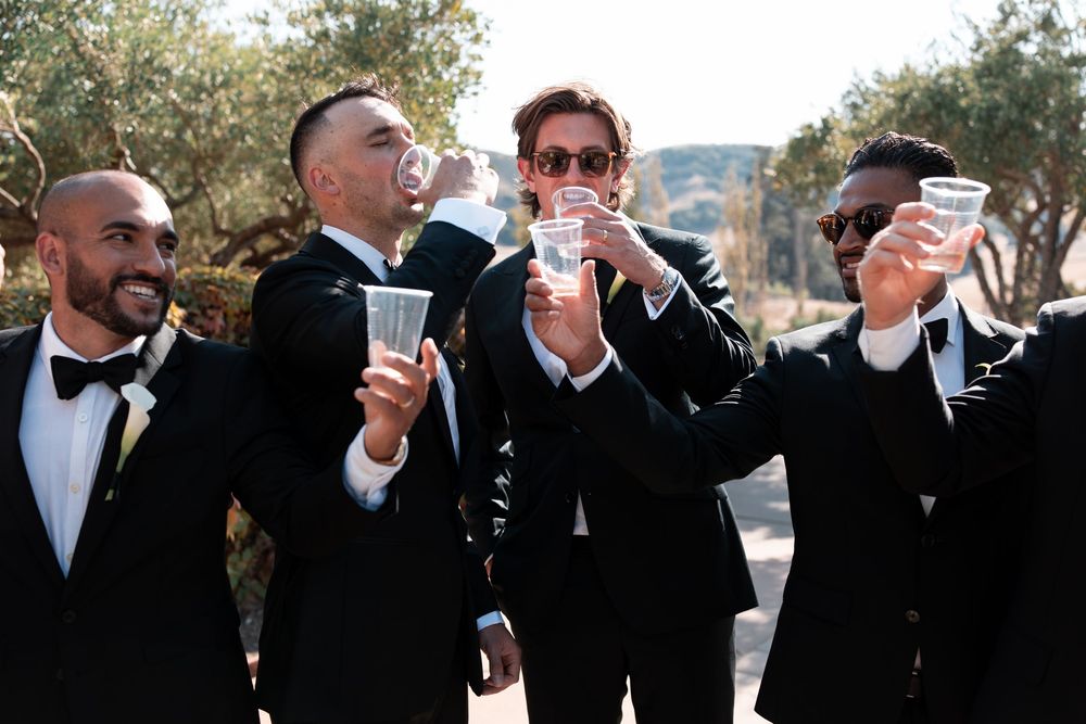 A sequence of wedding party members in black tuxedos raising their champagne glasses in celebration at an outdoor event.