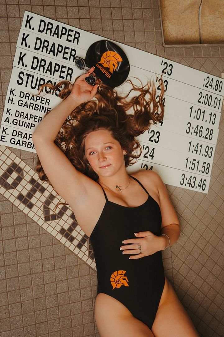Female swimmer poses with record board in black Spartan swimsuit and cap at indoor pool facility.