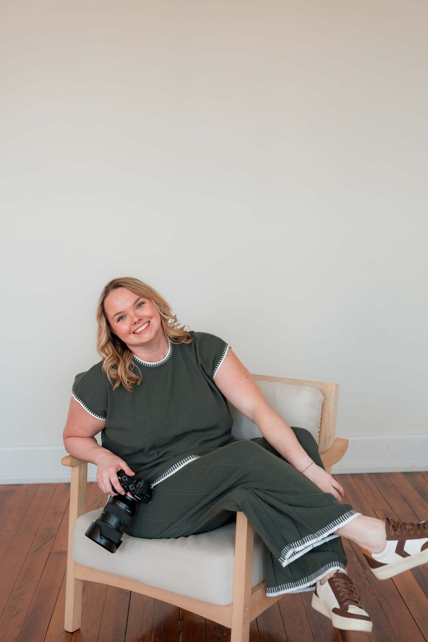 Person in olive green outfit relaxing in wooden chair while holding professional camera and wearing casual sneakers.