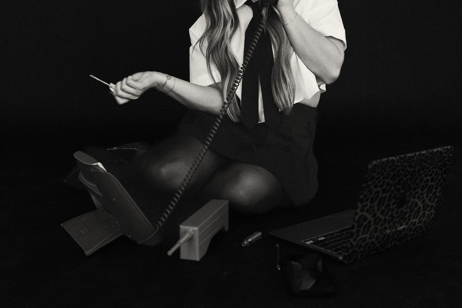 Black and white photo of woman in tie sitting on floor with telephone and laptop.