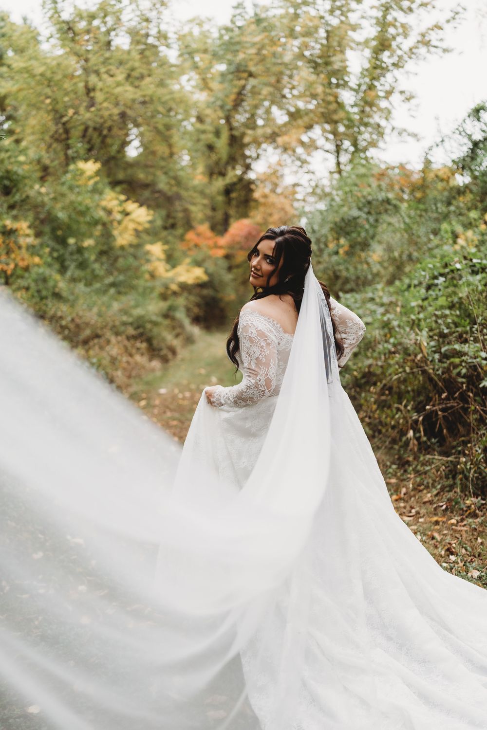 A flowing white wedding dress veil billows dramatically in the wind against an autumn forest backdrop.