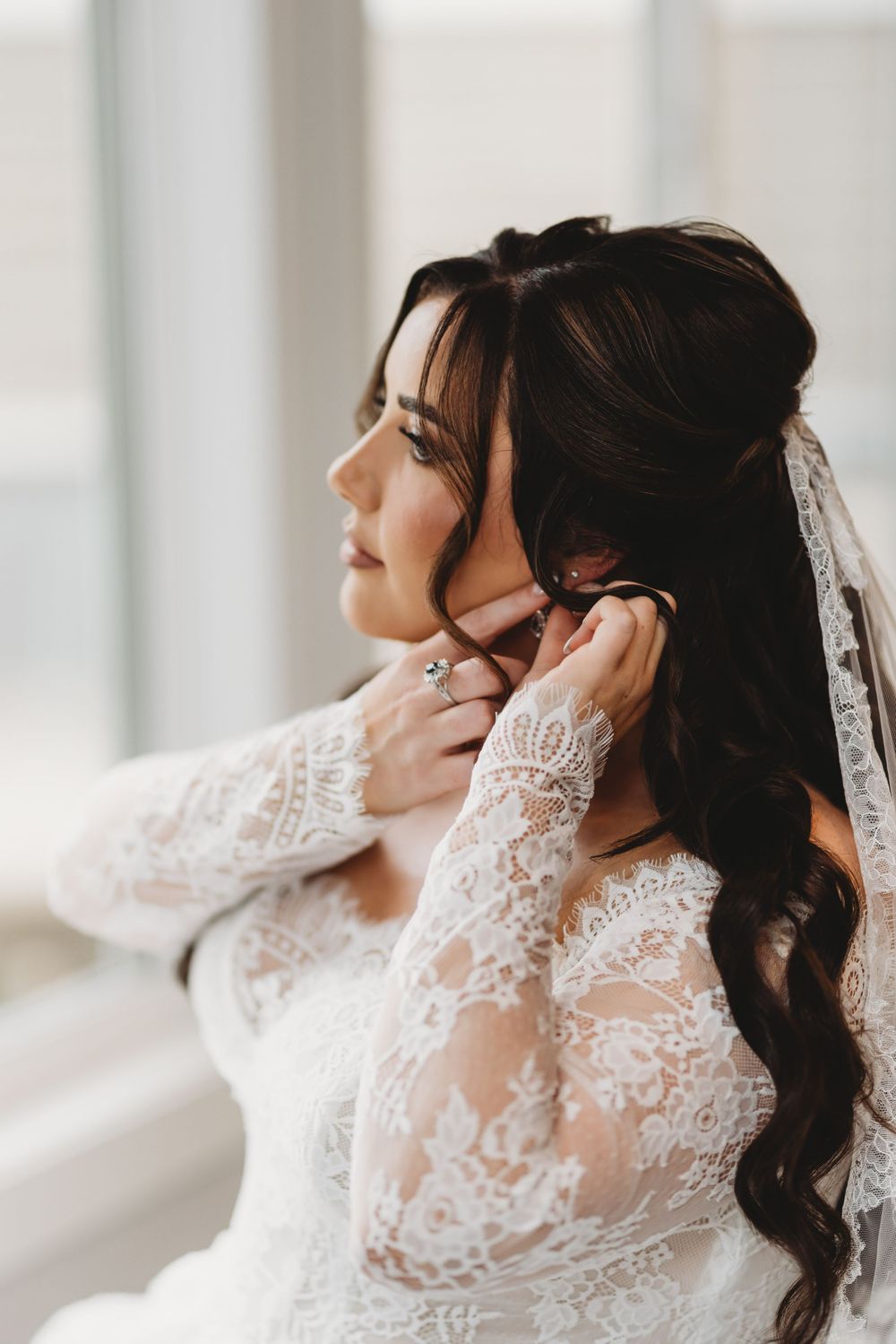 Bride in delicate lace wedding dress gazes thoughtfully to the side in a bright window-lit setting.