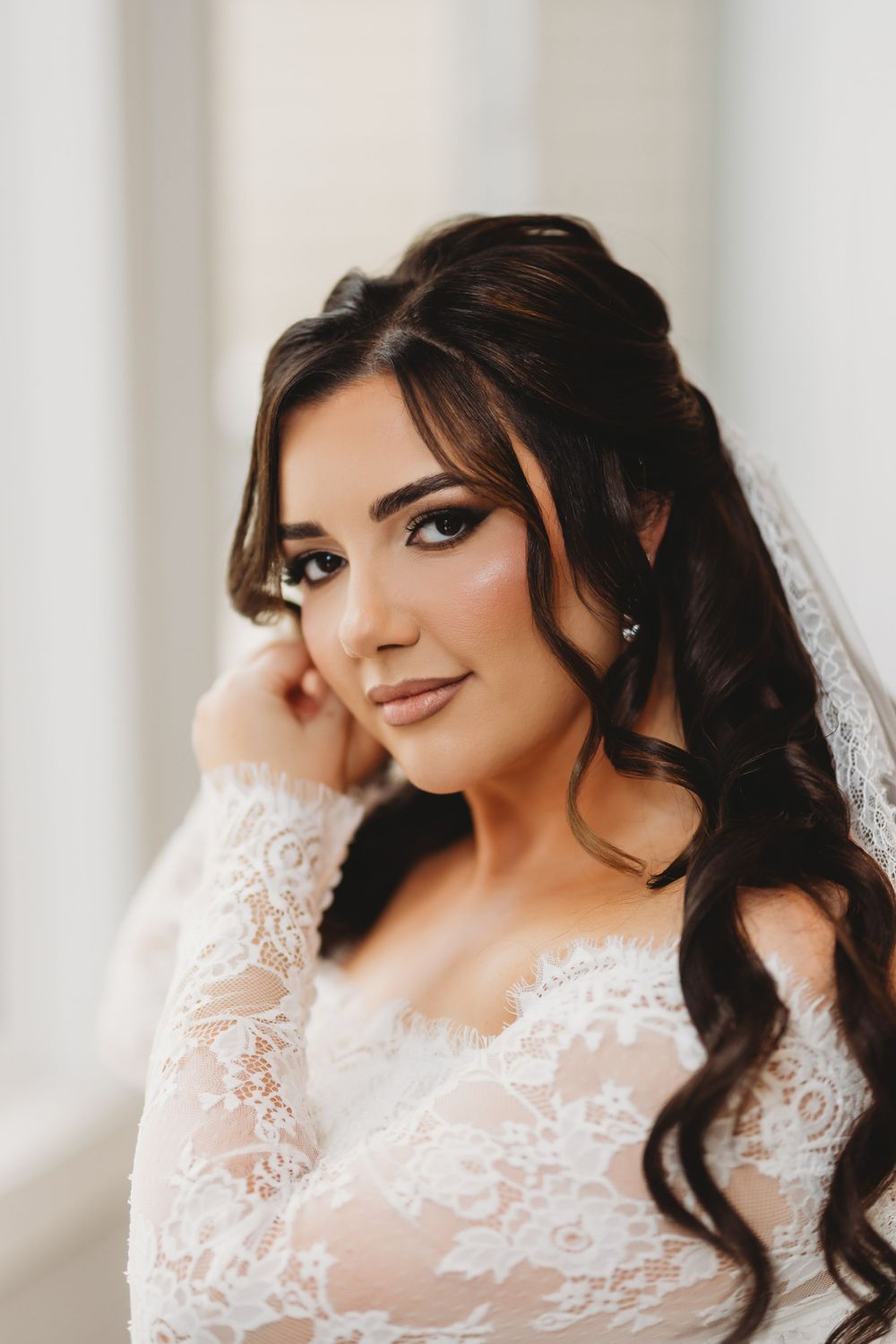 Beautiful bride in white lace wedding dress with long dark curls and delicate makeup poses for portrait.