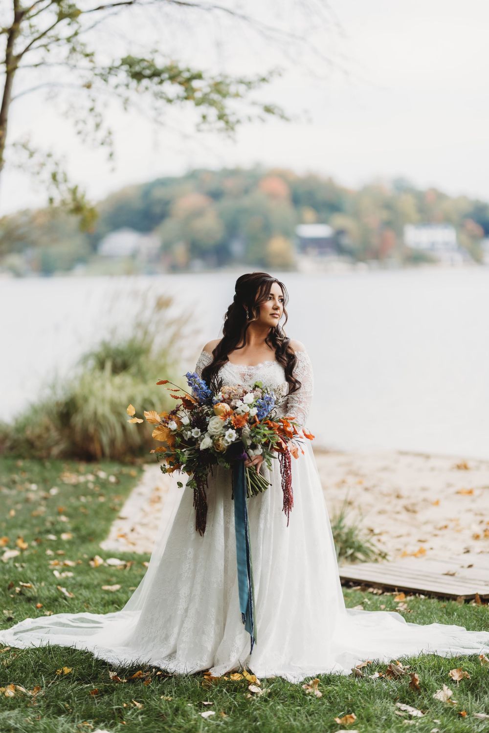 A bride in a flowing white gown holds a vibrant autumn bouquet while sitting by a lakeside on a fall day.