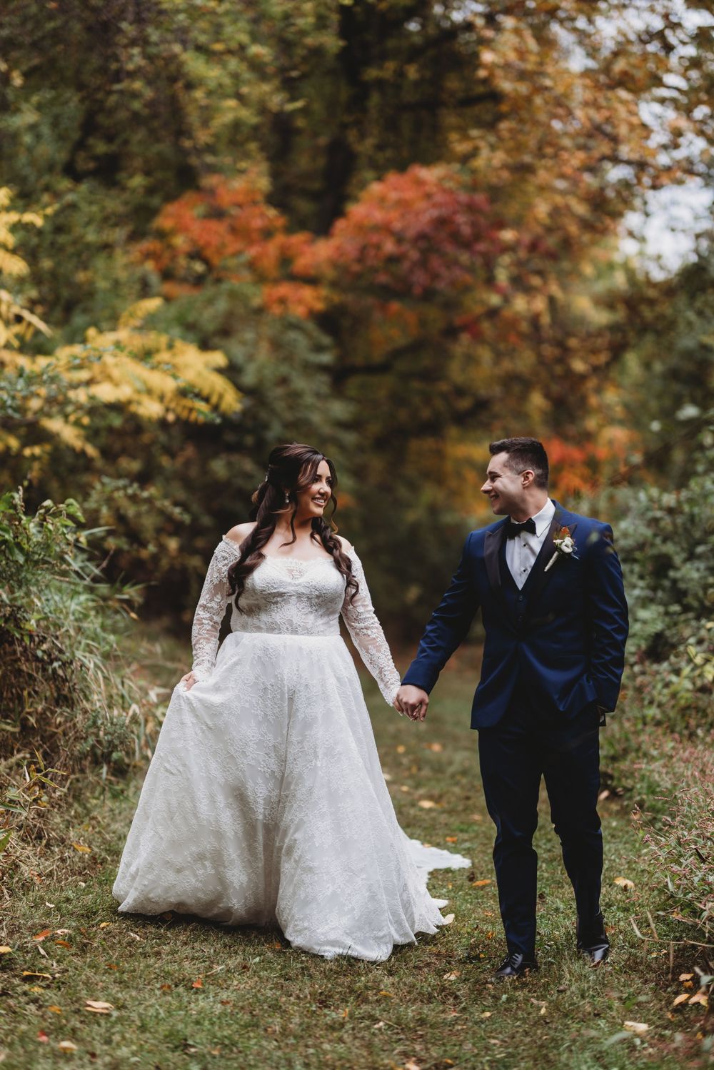 A couple in wedding attire walks together through a colorful autumn forest setting with vibrant fall foliage.