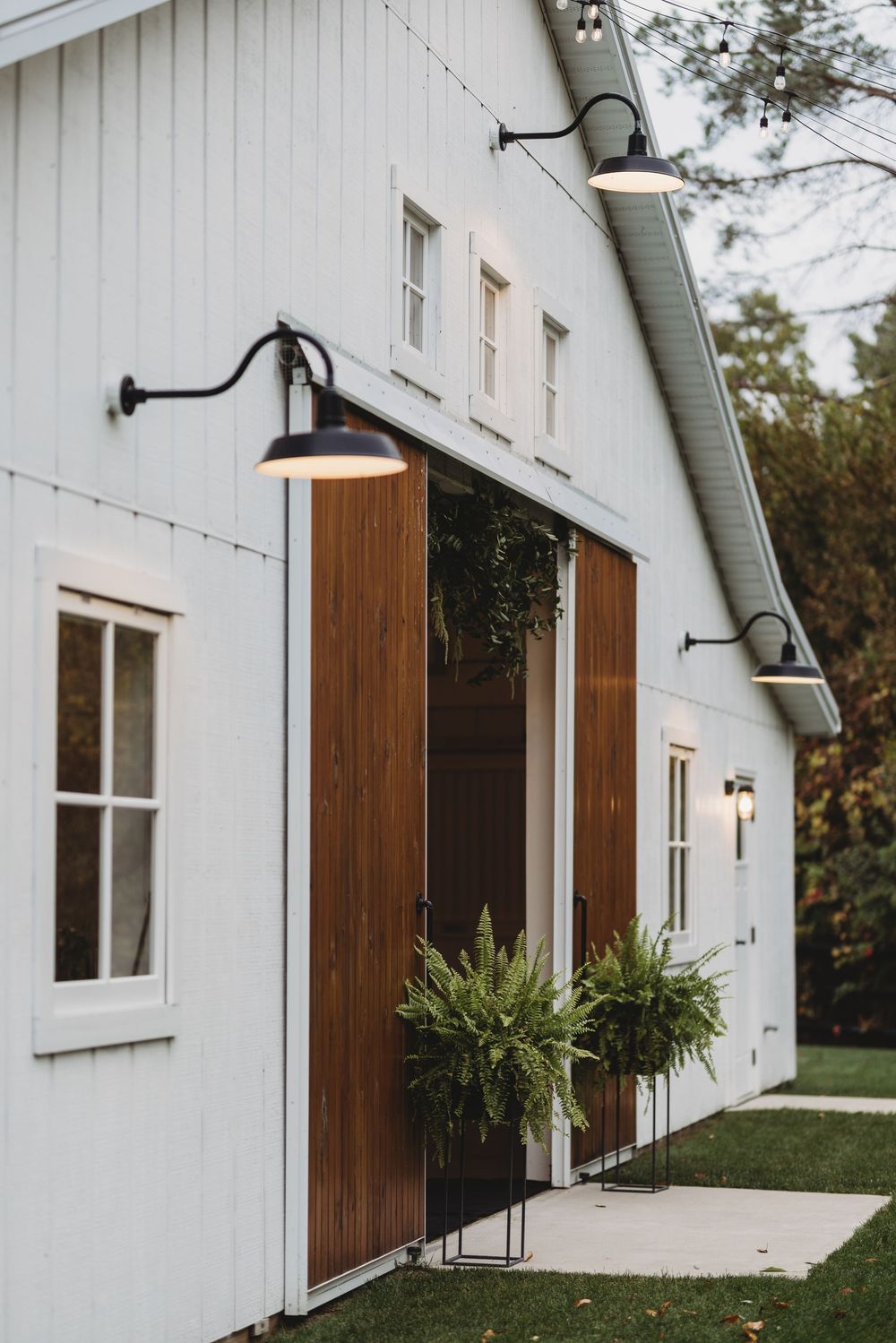 Modern farmhouse exterior features white siding, wooden barn doors, and black gooseneck lighting fixtures.