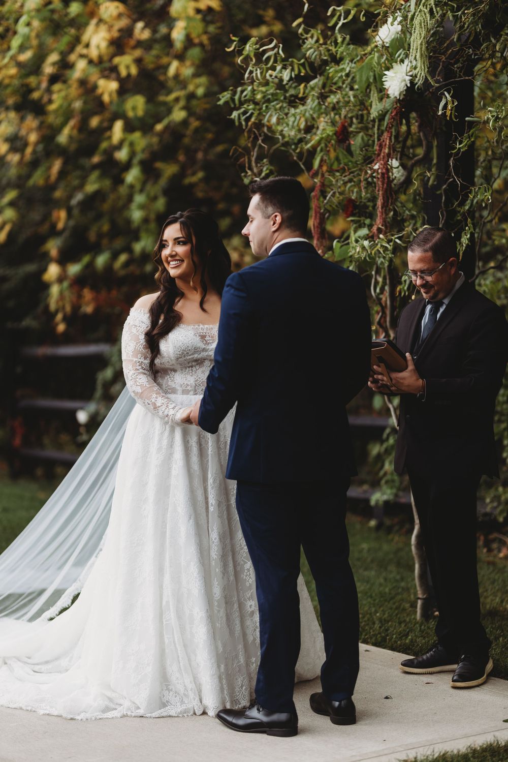 A romantic outdoor wedding ceremony with a couple in formal attire standing beneath trees in an autumn setting.