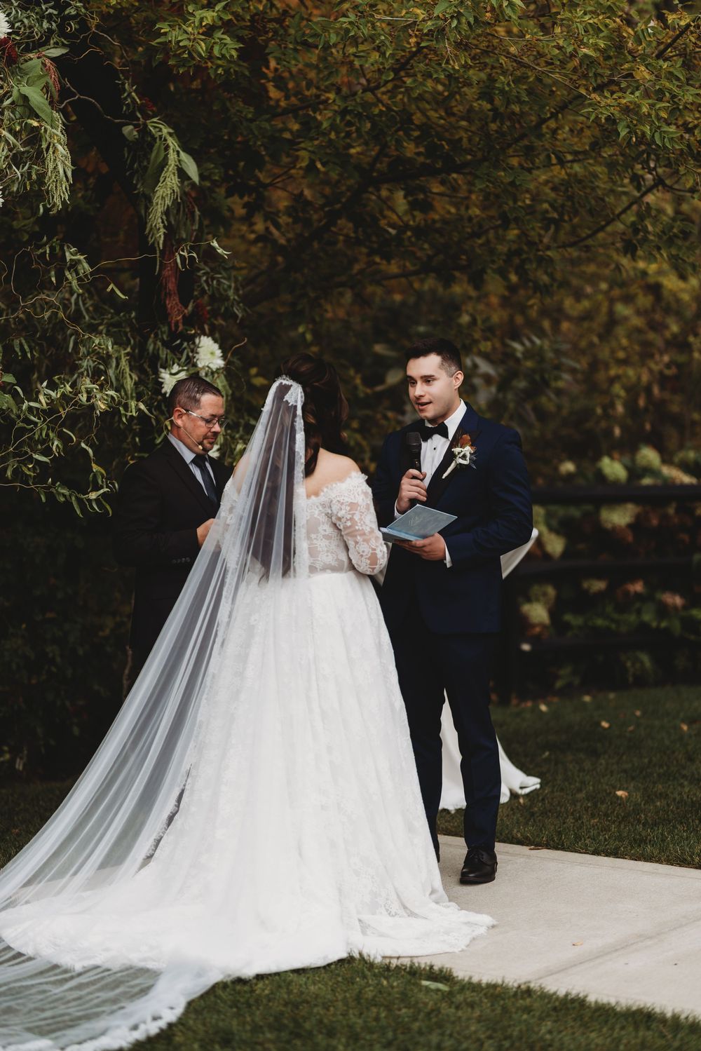 Newlywed couple reading vows outdoors during autumn wedding ceremony with long flowing white veil and navy suit.