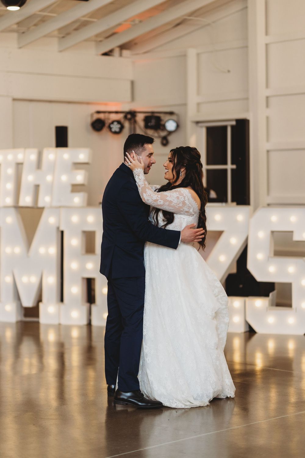 A couple shares their first dance in an elegant white reception hall with illuminated letters in the background.